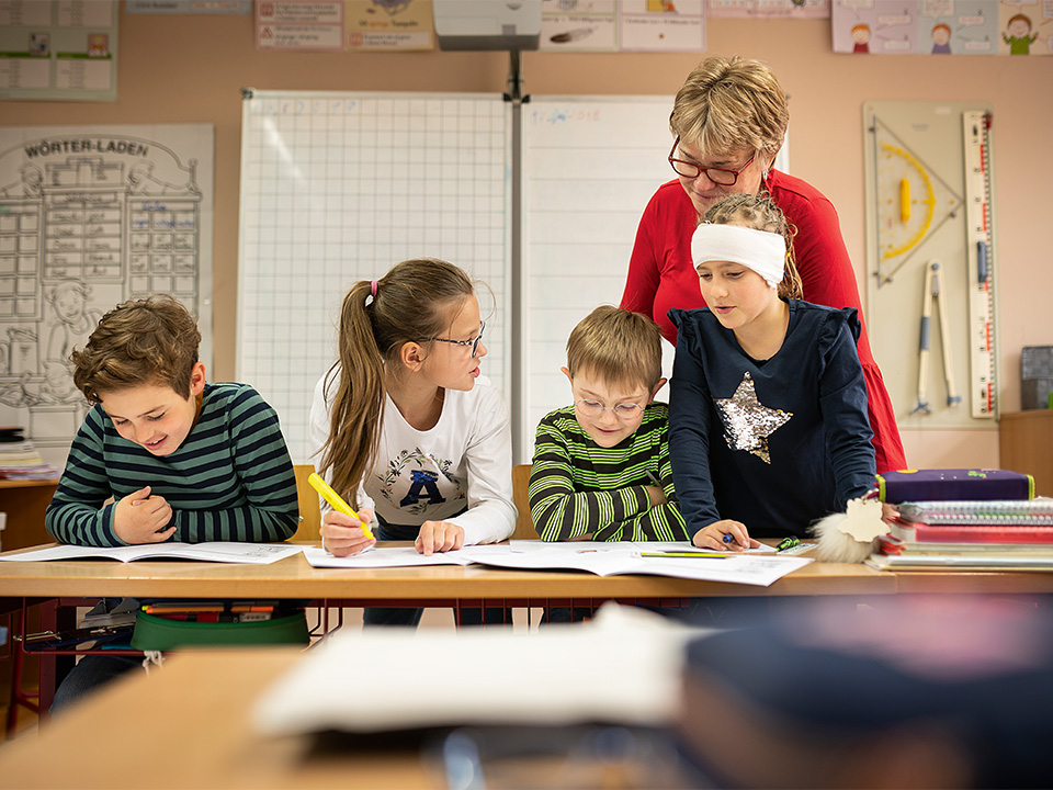 Vier Schülerinnen und Schüler sitzen am Tisch, eine Lehrerein steht hinter ihnen.